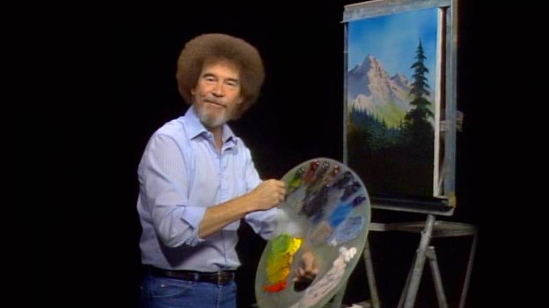 bob ross holds paint brush and palette in front of a painting